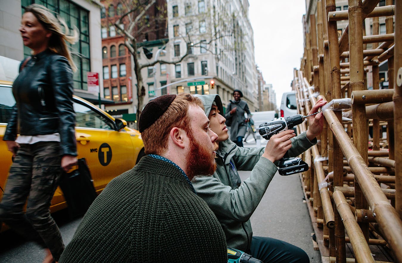 A young man in a hoodie drills into a wooden structure while another young man with a beard and a yarmulke watches
