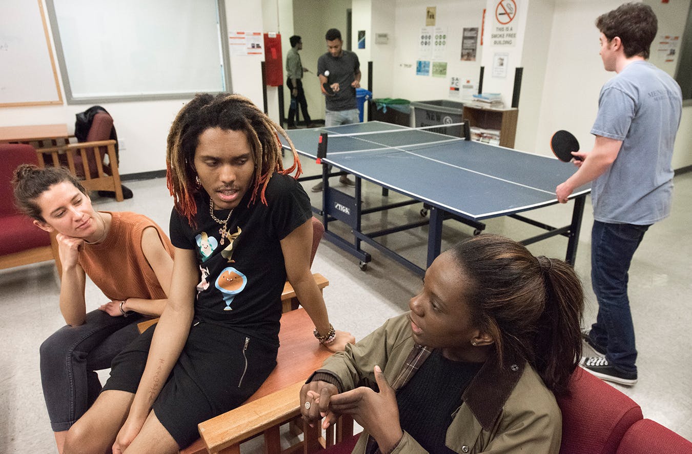 Three students talk in one of the student lounges while two other play ping pong.